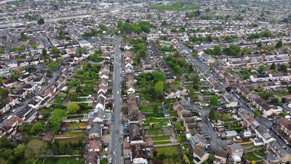 An aerial view of a residential neighbourhood showing rows of terraced houses and semi-detached homes, many with lush green gardens and well-maintained lawns. The gardens feature a variety of trees, s