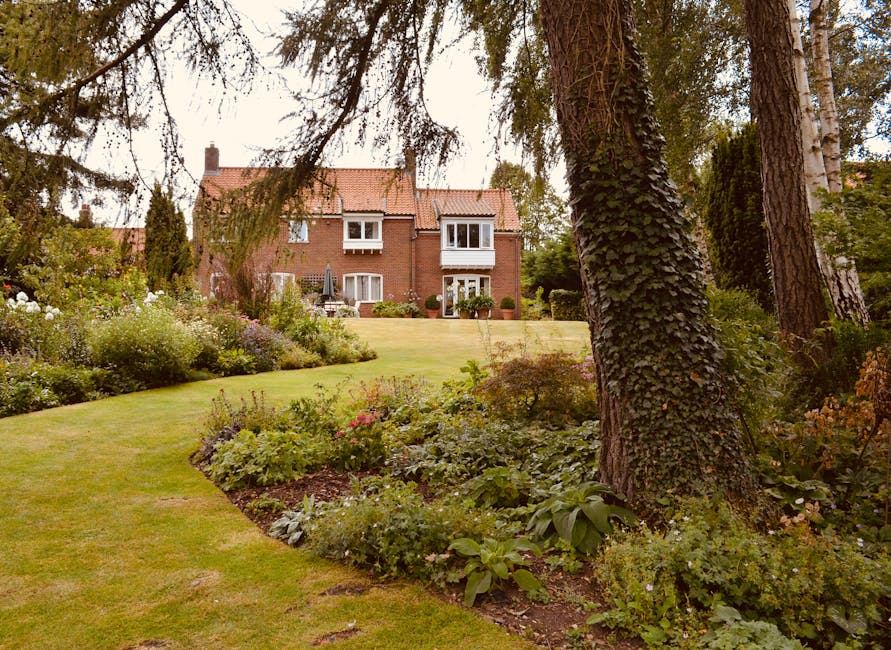 An aerial view of a residential neighbourhood showing rows of terraced houses and semi-detached homes, many with lush green gardens and well-maintained lawns. The gardens feature a variety of trees, s