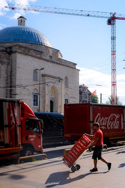 The image depicts a construction crane towering above a historic building with a large blue dome and light-colored stone walls. In the foreground, a man dressed in a red shirt and black shorts is push